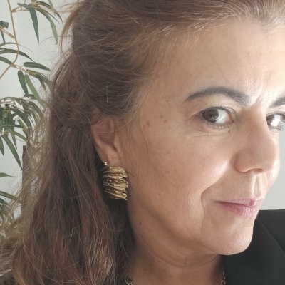 Brinco dourado texturizado usado por mulher com cabelo castanho e planta verde ao fundo