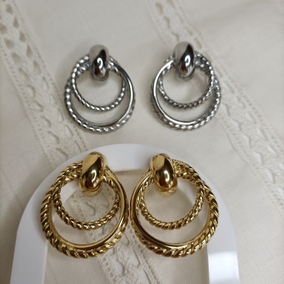 Dois pares de brincos mock de argolas entrelaçadas, um prata e outro dourado, sobre tecido branco com rendilhado