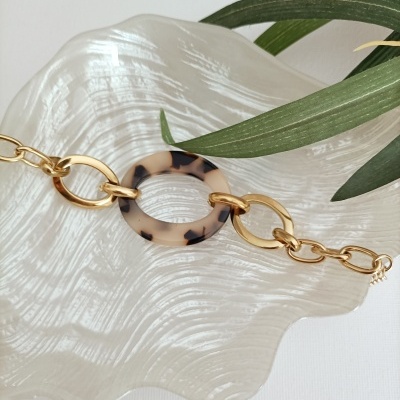 Pulseira dourada com círculo em padrão tartaruga sobre prato branco texturizado e folhas verdes