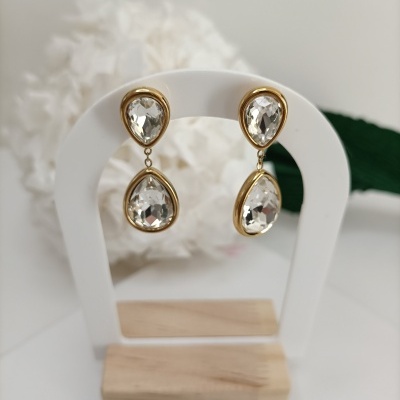 Brincos dourados com pedras transparentes em forma de gota em suporte branco e madeira com fundo com flor branca