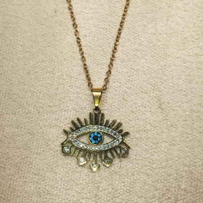 Colar dourado com pendente de olho com pedras azuis e transparentes sobre tecido bege