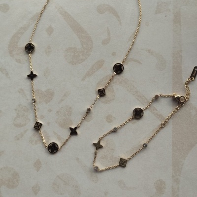 Colar e pulseira dourados com pendentes geométricos e florais, sobre superfície bege com padrão.