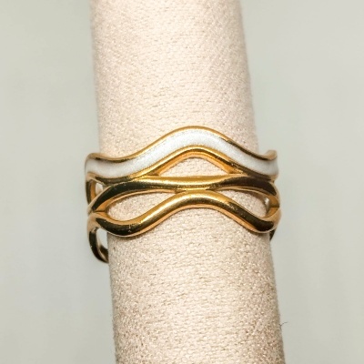 Anel com três bandas metálicas entrelaçadas, duas douradas e uma prateada, num suporte creme
