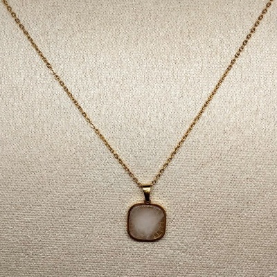 Colar dourado com pendente quadrado de pedra branca translúcida em fundo bege.