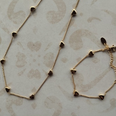 Colar e pulseira dourados com pequenos corações sobre fundo claro com padrão