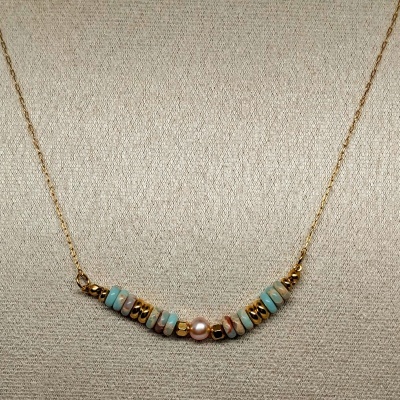 Colar dourado com discos azuis, dourados e pérola central sobre fundo bege.