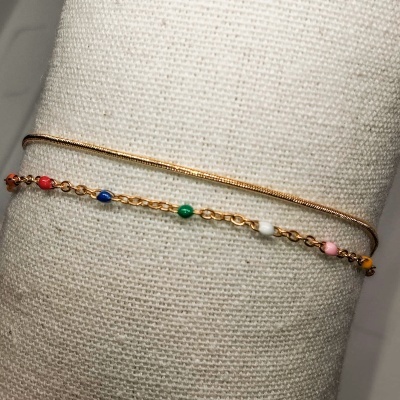 Dois braceletes dourados finos sobre almofada bege, um com pérolas coloridas e outro liso.