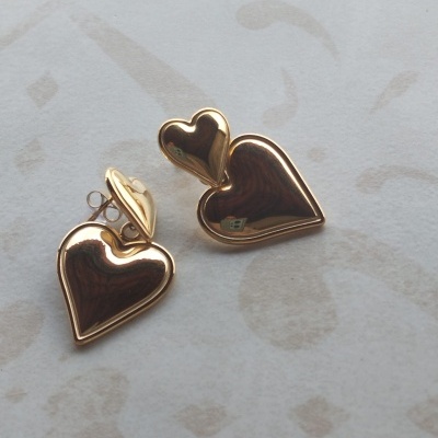 Brincos dourados em forma de coração com dois tamanhos, fundo claro com padrões abstratos.