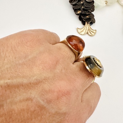 Mão a usar dois anéis dourados com pedras castanha e verde oliva