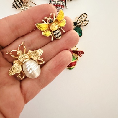 Broches em forma de abelha metálica dourada com detalhes coloridos, mostrando várias peças sobre a mão.