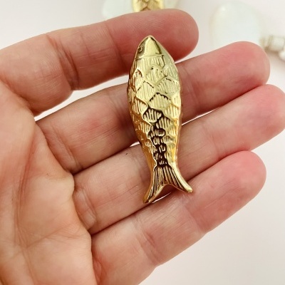 Peça decorativa dourada em forma de peixe segurada por uma mão com pedras brancas ao fundo