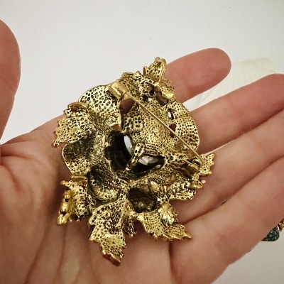 Broche dourado decorativo em forma de folhas, visto pela parte de trás, segurado numa mão.