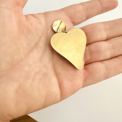 Brinco dourado formato coração segurado numa mão.
