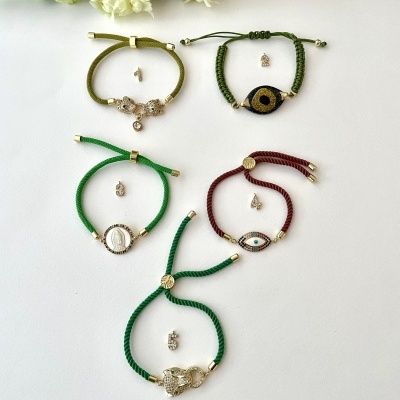 Cinco pulseiras coloridas com pendentes metálicos dourados em fundo branco e flores ao fundo