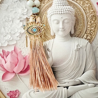Estátua branca de Buda com flores ao fundo e pendente dourado com franjas e olho turco.