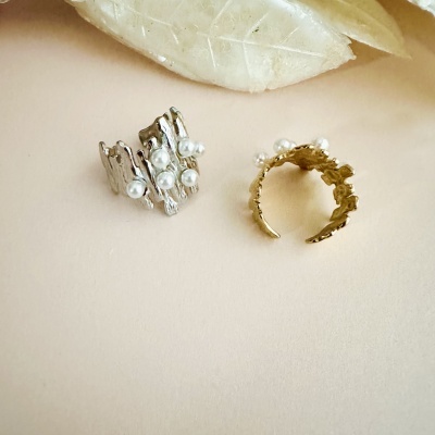 Dois anéis abertos com pérolas falsas, um prateado e outro dourado, sobre superfície clara com folha decorativa.