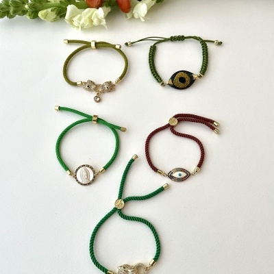 Cinco pulseiras coloridas de cordão com detalhes dourados e amuletos variados, em fundo branco, com flores ao fundo.