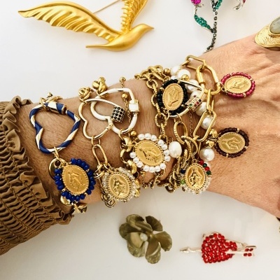 Braceletes dourados com medalhas e pedras vistosas numa mão com manga castanha, com broches coloridos ao fundo