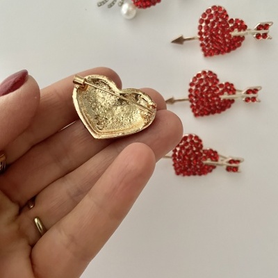 Broches em forma de coração com flecha, quatro com pedras vermelhas e um mostrado pelo verso em dourado, sobre fundo branco.