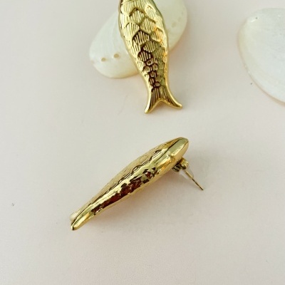 Brincos dourados em formato de peixe com textura de escamas sobre superfície clara