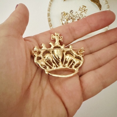 Broche dourado em forma de coroa segurado numa mão sobre fundo branco