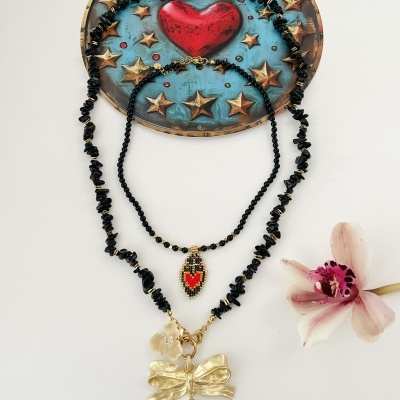 Três colares negros com pingentes coloridos sobre fundo branco, prato decorativo azul com estrelas douradas e coração vermelho e orquídea rosa clara