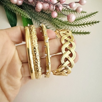 Conjunto de quatro pulseiras douradas metálicas com texturas variadas numa mão com fundo de folhas e bagas