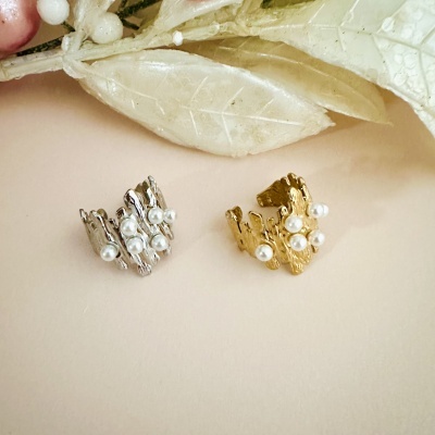 Dois anéis decorados com pérolas, um prateado e outro dourado, sobre fundo bege com folhas artificiais.