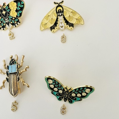 Quatro broches esmaltados de insetos com números em strass pendurados, sobre fundo branco.