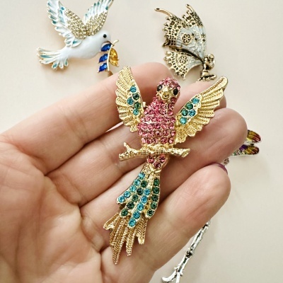 Broche dourado com pedras coloridas em forma de pássaro, na mão, com outros broches ao fundo.