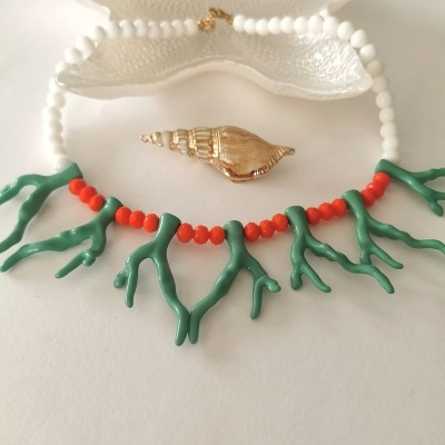 Colar com contas brancas e vermelhas, pendentes verdes em forma de ramos, em cima de concha branca decorativa e concha dourada ao lado.