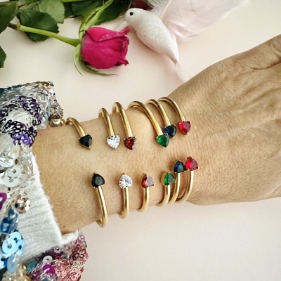 Pulseiras douradas com pedras de coração coloridas no pulso junto a uma rosa vermelha e pássaro branco