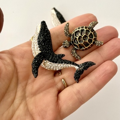 Broches em forma de baleia e tartaruga com pedraria, em mão sobre fundo branco
