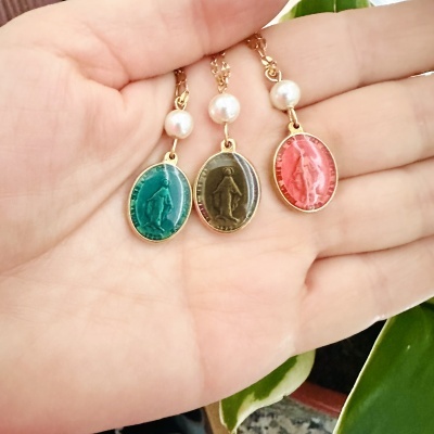 Três colares com medalhas ovais coloridas penduradas numa mão, com pérolas brancas e correntes douradas.