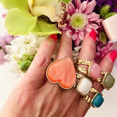 Mão com anéis de pedras coloridas e unhas vermelhas em fundo de flores.