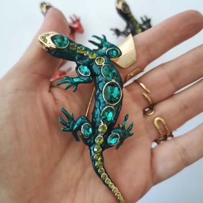 Broche de lagarto verde e dourado com pedras brilhantes numa mão.