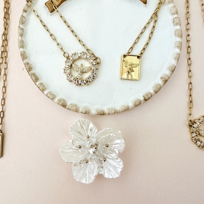 Colares dourados com pingentes delicados e acessório floral branco numa superfície rodada branca.