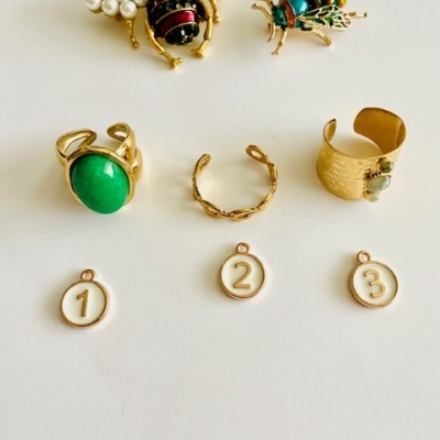 Acessórios dourados incluindo pins com forma de abelha, três anéis e três medalhas numeradas sobre fundo branco