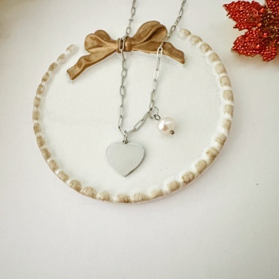 Colar prateado com pendente em forma de coração e pérola numa base branca decorada com fita dourada e ornamento vermelho.