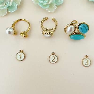 Anéis dourados com pérolas e pedras azuis junto a flores brancas decorativas e etiquetas numeradas