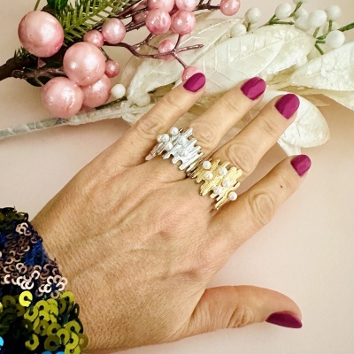 Mão com dois anéis texturizados em prata e dourado com pérolas, unhas roxas, fundo com ramos decorativos.