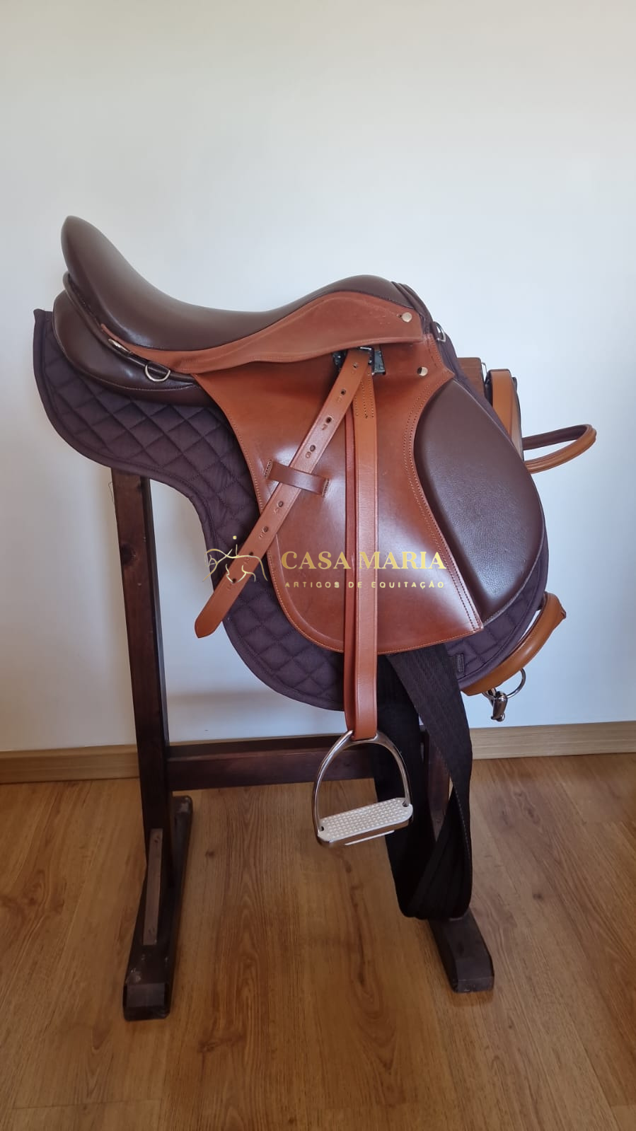 Sela de equitação em couro castanho sobre almofada castanha escura em suporte de madeira