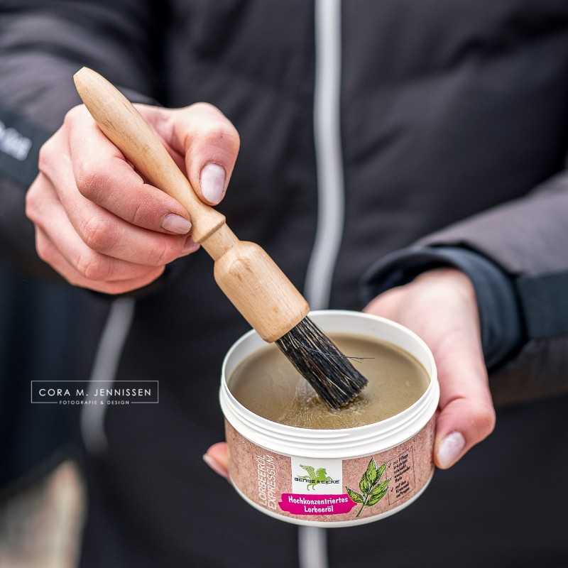 Pincel de cabo de madeira sendo mergulhado em produto dentro de lata branca com etiqueta verde e bege, segurado por uma mão com casaco preto.