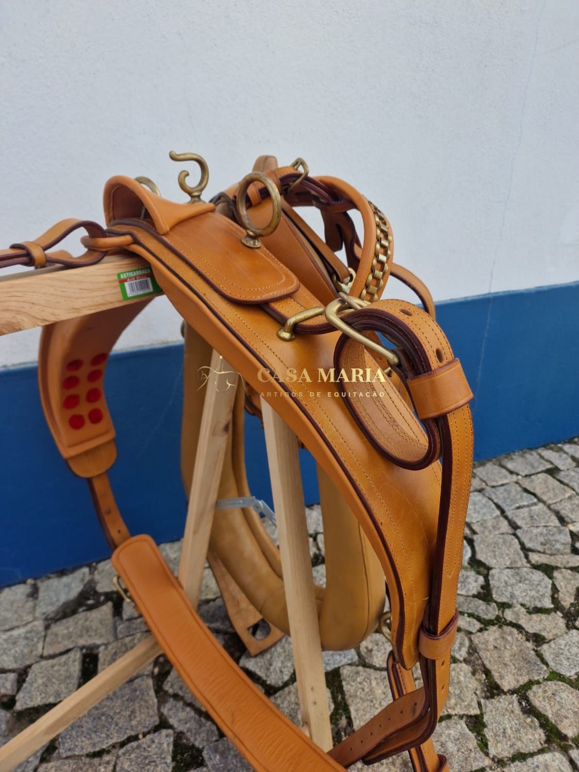 Selaria de montar em couro castanho claro com argolas e fivelas douradas pendurada em estrutura de madeira