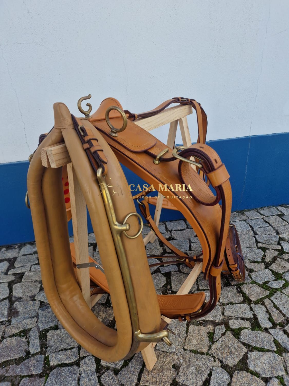 Sela de equitação em couro castanho claro com ferragens douradas num suporte de madeira sobre calçada empedrada.