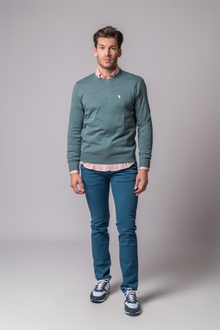 Homem com camisola verde, camisa xadrez, calças azul e ténis desportivos no fundo cinzento