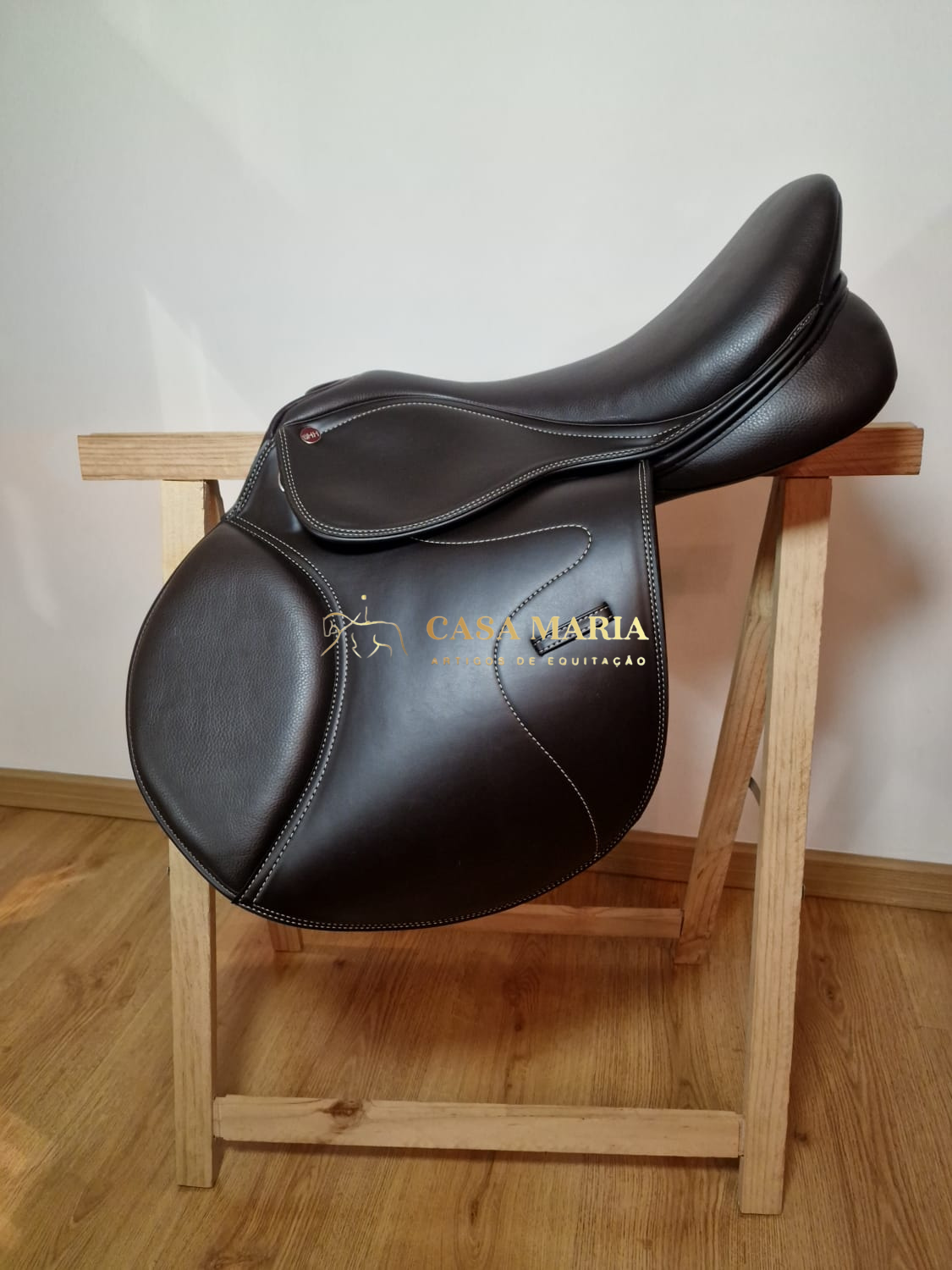 Sela de equitação em couro castanho escuro sobre suporte de madeira clara com logo CASA MARIA