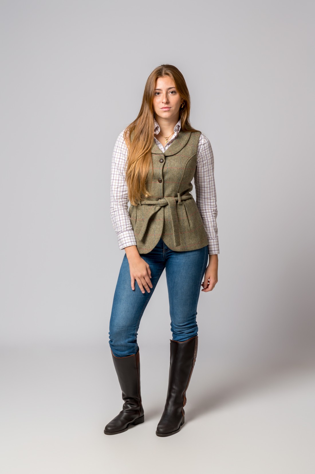 Mulher com camisa às riscas, colete bege com cinto, jeans azul e botas castanho-escuras, fundo cinzento