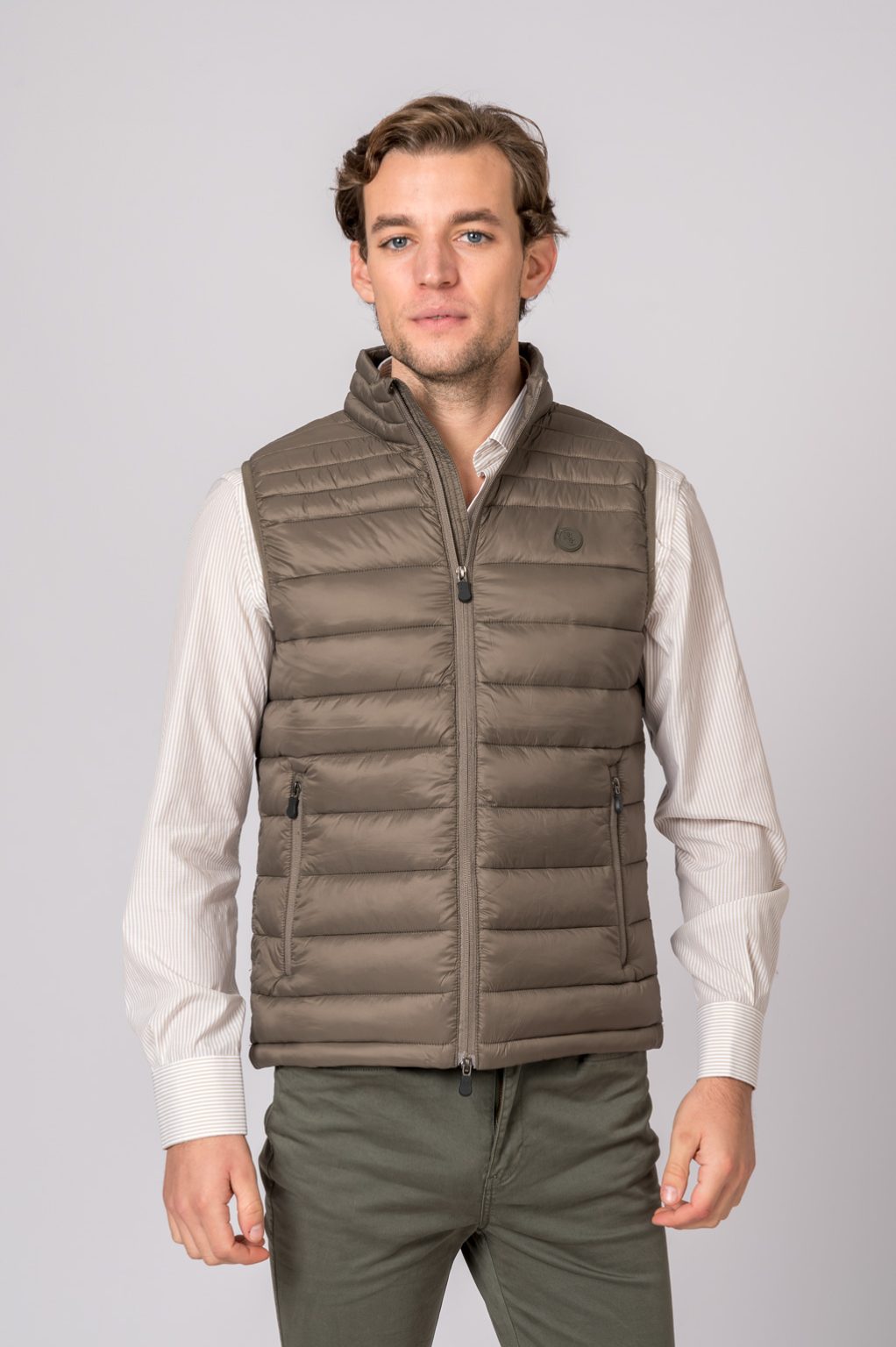 Colete acolchoado castanho claro para homem, com fecho e bolsos laterais, usado sobre camisa creme de riscas finas