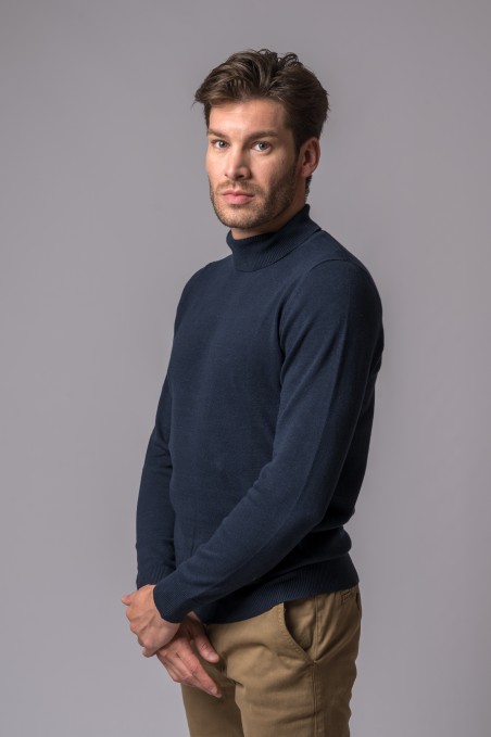 Homem com camisola de gola alta azul escura e calças bege em fundo cinzento claro.